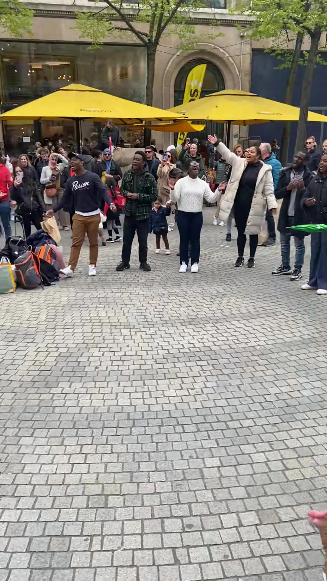 A wide ring of people on the Meir in Antwerp around a woman singing, café parasols behind.